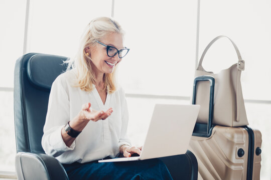 Mature woman using a laptop at an airport, waiting with luggage for her next exciting journey to explore new destinations - Powered by Adobe