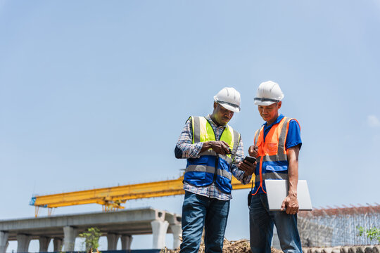 Engineers in hard hats and vests looking at a smartphone, Construction workers reviewing project plans on construction site, Civil engineers working at a new road construction site