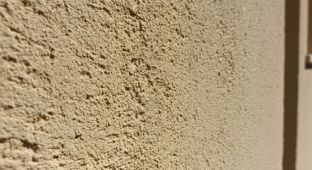 Close-up reveals the intricate texture of a beige stucco wall, where light plays to enhance the rough, granular surface and deep shadows, creating a warm, organic and tactile visual experience.
