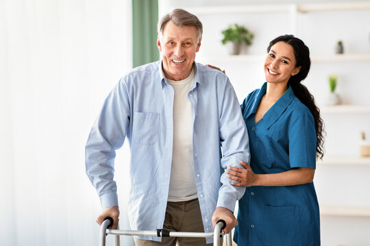 Portrait of happy elderly man using walker and lovely young nurse helping him at retirement home. Mature male petient with caregiver going for walk at seniors centre. Professional eldercare service