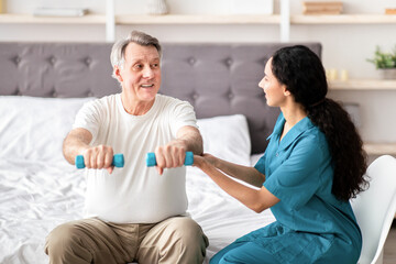 Young rehabilitation therapist assisting senior male patient with strength exercises with dumbbells...