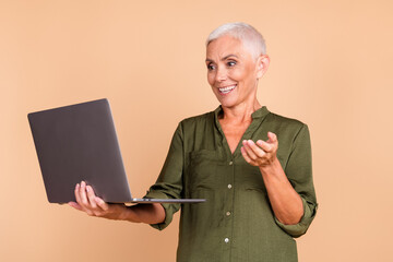 Confident senior woman with short white hair holding a laptop, gesturing happily against a beige...