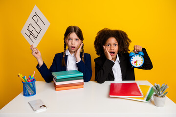 Two young schoolgirls showing stress at their desk with books and a clock against a bright yellow background