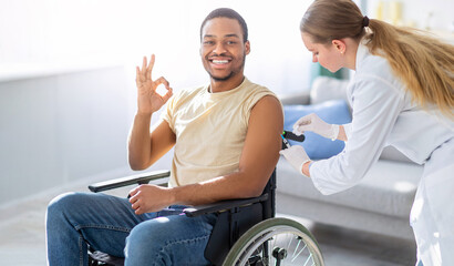 Immunizing homebound patients. Young doctor giving injection against flu to young with disability man at home. Black guy in wheelchair showing ok gesture, receiving vaccination