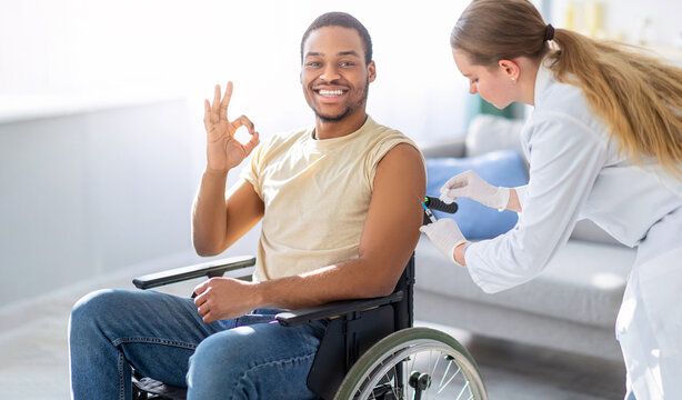 Immunizing homebound patients. Young doctor giving injection against flu to young with disability man at home. Black guy in wheelchair showing ok gesture, receiving vaccination