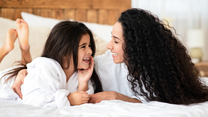 Beautiful young mother and little daughter in bathrobes laying on bed at home, hugging and...