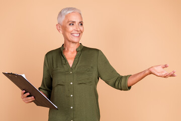 Elegant senior woman in casual khaki shirt smiling and gesturing while holding black clipboard against warm beige background