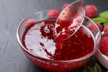 Taking sweet raspberry jam with spoon from glass bowl on gray textured table, closeup