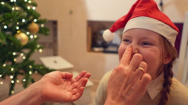 Closeup view of woman applying cosmetic on girl face sitting in a room near Christmas tree. Slow motion of mother doing makeup to her teenage daughter with sparkling shimmer. Beauty and fashion theme.