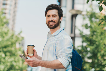 Handsome man enjoying coffee outdoors while checking his phone and smiling at the camera in a sunny urban setting.