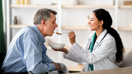 Fototapeta premium Caregiver feeding elderly man in wheelchair at home. Older male patient with restrained movement after stroke eating with caregiver's help. Medical assistance for senior people with partial paralysis