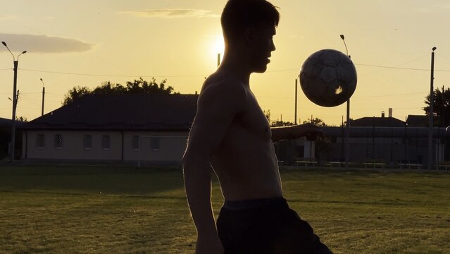 Young man juggling soccer ball on stadium at sunset. Professional footballer kicking ball at green field. Sportsman practicing tricks at meadow with sunlight at background. Freestyle football - Powered by Adobe
