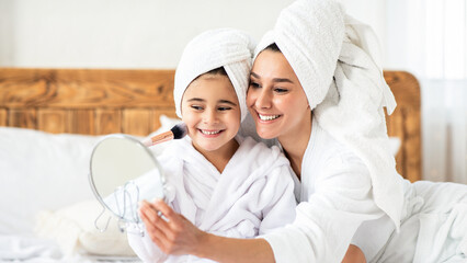Mom putting make up on her cute little daughter, tickling her with make up brush. Cheerful mother in bathrobe with hair wrapped in towel applying foundation on her kid cheeks, bedroom interior