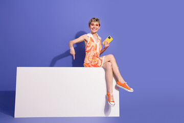 Youthful woman in a vibrant summer outfit seated against a colorful background holding a phone and...