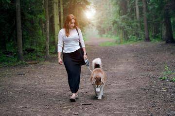 A woman walks her beautiful husky dog on a path through a lush green forest. The pair are enjoying a peaceful stroll surrounded by nature and greenery © aapsky