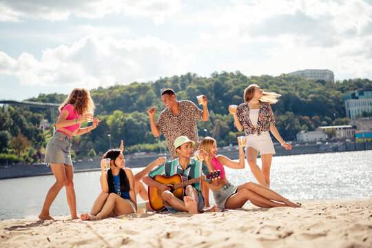 Group of friends enjoying a summer day on the beach with guitar playing and laughter, celebrating life together by the river