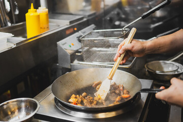 Chef stir-frying meat and vegetables in a wok on a stovetop in a professional kitchen, with cooking tools and condiments visible in the background.