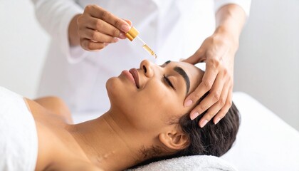 Fototapeta premium Closeup of beautician’s hand using dropper to apply facial serum on young woman’s forehead during spa procedure. Skincare therapy for moisture balance and radiant complexion.