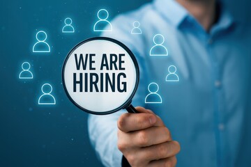 A person holds a magnifying glass highlighting the message "we are hiring" surrounded by translucent people icons, conveying a recruitment concept.