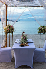Wedding arch on coast. Romantic beachside dining setup featuring beautifully adorned table with floral arrangements, soft lighting and serene ocean backdrop, creating intimate atmosphere. Vertical