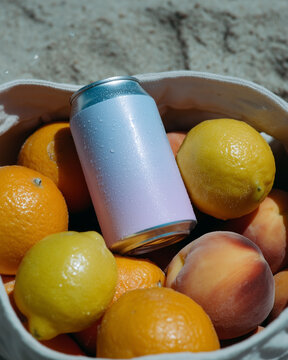 A soda can lying on the beach in a bag with peaches, lemons, and oranges. Refreshing chilled drinks.
