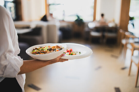 Waiter in white shirt serving two plates with gourmet food and fresh salad in a bright, modern restaurant interior. Selective focus on dishes