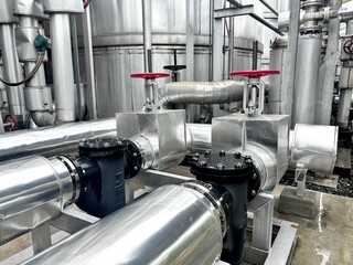 The image shows a messy, industrial indoor space with a complex network of pipes, valves, and mechanical equipment. A small staircase with a yellow railing and a danger sign leads up to a tank.