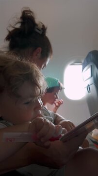 Mother holding baby with teether and book while child sits by airplane window, family travel scene showing close interaction during flight