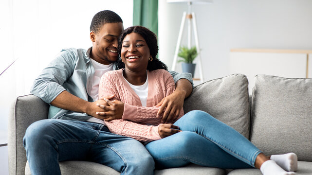 Joyful african american young couple in casual sitting on couch, hugging and laughing, living room interior, copy space. Happy black family spending cold winter day together at home, cuddling