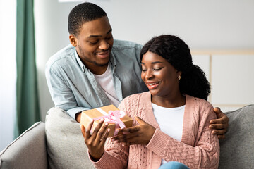 Birthday, anniversary, international women's day celebration concept. Romantic black guy giving present his happy girlfriend, african american young family having marriage anniversary, home interior