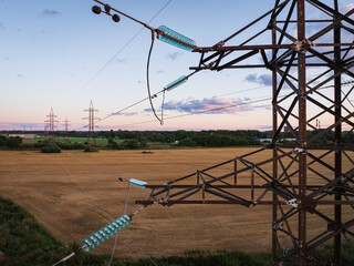 Close-up of a high-voltage electricity pylon and transmission power lines with glass insulators in a rural field at sunset.