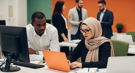 Two colleagues are working at separate desks in a modern office, with a woman in a hijab typing on an orange laptop and a man looking at a desktop monitor, while other blurred coworkers stand in th..