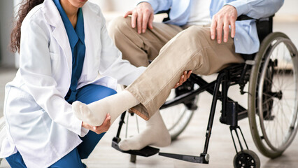 Cropped view of young doctor checking disabled senior patient's leg during medical visit at home....