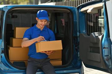 Smiling professional courier signing for package delivery, completing during parcel drop-off at...