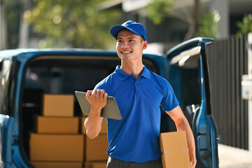 Portrait of a confident courier in a blue uniform holding a cardboard box and tablet, standing...