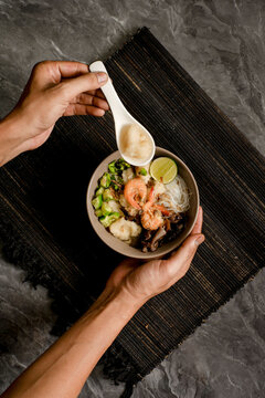 Top view of tekwan, Palembang fish ball soup, with shrimp, vermicelli, mushrooms and lime. Hands hold bowl and spoon. Perfect for menu design, blog headers, and Asian cuisine promos.