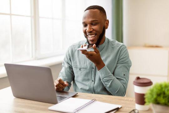 African Businessman With Smartphone Using Voice Assistant App Or Loudspeaker Talking Holding Phone Near Mouth Sitting At Workplace In Modern Office. IOT, Business Applications And Voice Search - Powered by Adobe