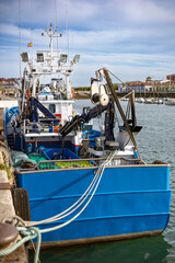A blue commercial fishing trawler is docked at a pier in a busy harbor. The vessel is equipped with a hydraulic multifunctional manipulator and other fishing gear.