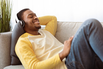 Obraz premium Happy african american guy enjoying music, using headset, side view. Smiling young black man with closed eyes listening to music while chilling alone at home, using brand new headphones