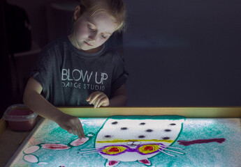 Beautiful little girl draws with sand on a light square, art therapy, sand animation, creative leisure for children, selective focus	