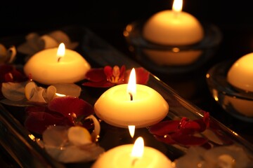 Burning candles and orchid flowers in glass container of water against black background, closeup
