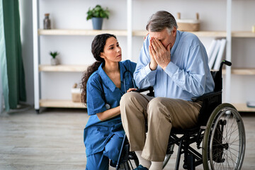 Naklejka premium Depressed elderly male in wheelchair crying, young nurse consoling him at retirement home. Unhappy impaired man feeling lonely and sad, millennial caregiver offering him support