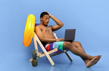 Surprised African American guy in swimwear sitting in lounge chair with laptop, looking at screen...