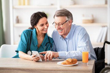 Disabled senior man and young nurse using smartphone together, browsing web or watching movie indoors. Handicapped elderly patient and his caregiver checking social media on mobile device