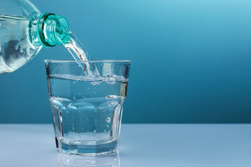 Pouring refreshing soda water from bottle into glass at white mirror table against light blue...