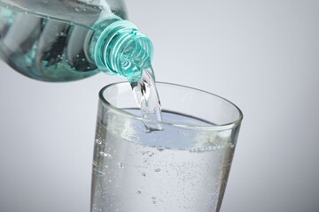 Pouring refreshing soda water from bottle into glass at table against light grey gradient...