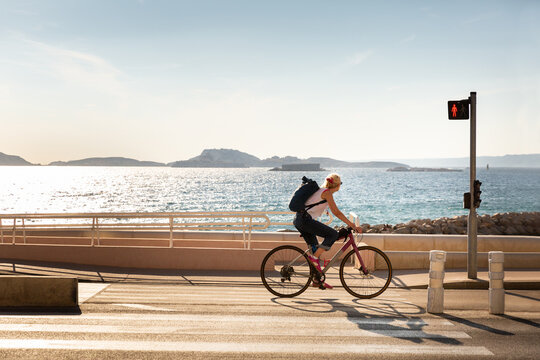 Femme sur un v&eacute;lo sur la piste cyclable de la corniche Kennedy &agrave; Marseille avec la mer et les &icirc;les en arri&egrave;re-plan, la femme est vue de profil sur son v&eacute;lo, elle regarde la mer et porte un sac &agrave; dos