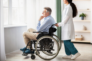 Side view of young doctor helping elderly disabled man in wheelchair, indoors. Nurse taking care of...