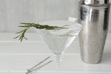 Martini cocktail with rosemary in glass and shaker on white wooden table, closeup