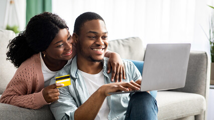 Happy african american couple with credit card and laptop sitting on floor in living room, shopping...